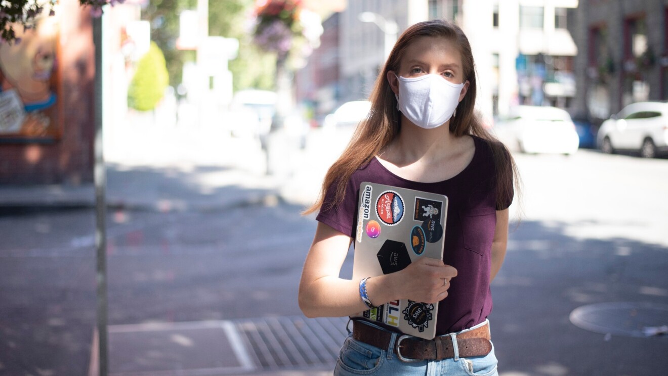 A woman stands on the street with a mask on, looking at the camera. She is holding a laptop with stickers related to Amazon and AWS.