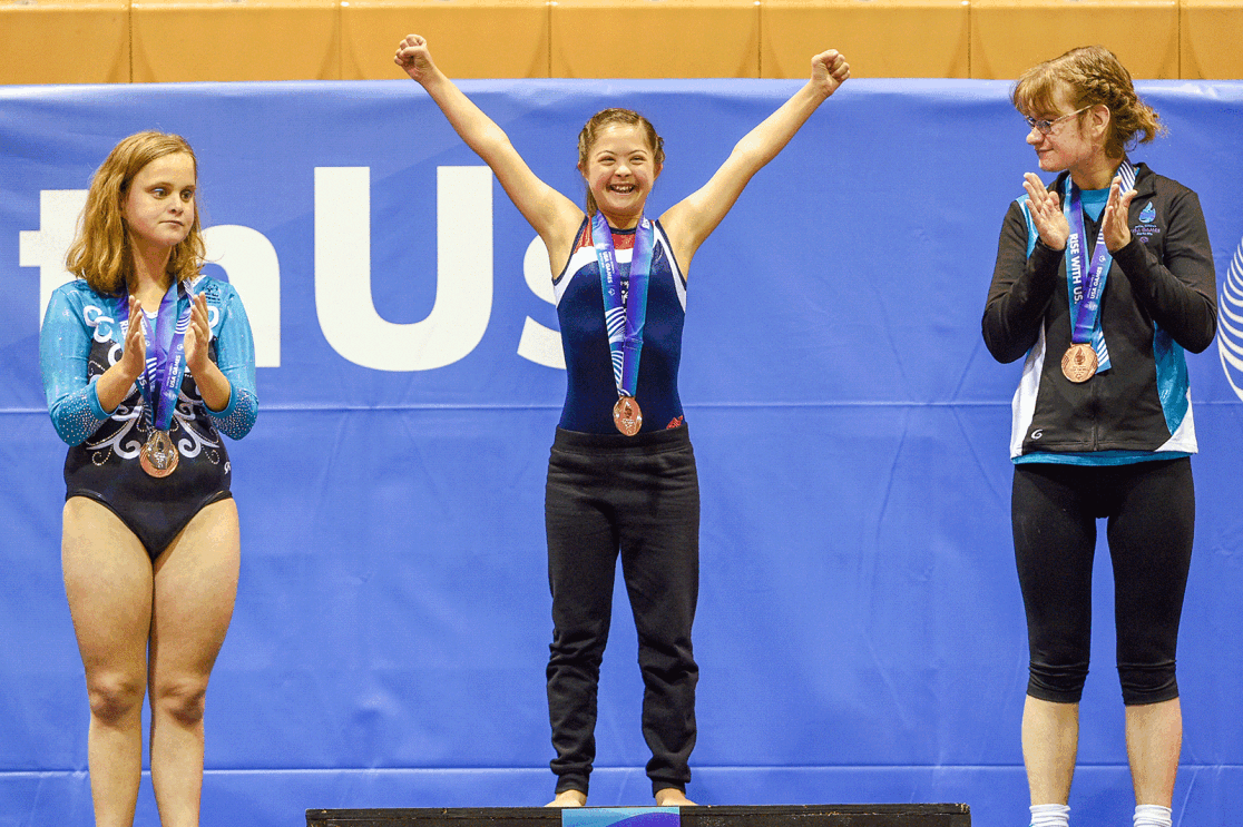An image of three athletes standing in a podium. The center of the image includes a girl with her arms up wearing a medal. There are two girls, one on each side, wearing medals and clapping. 