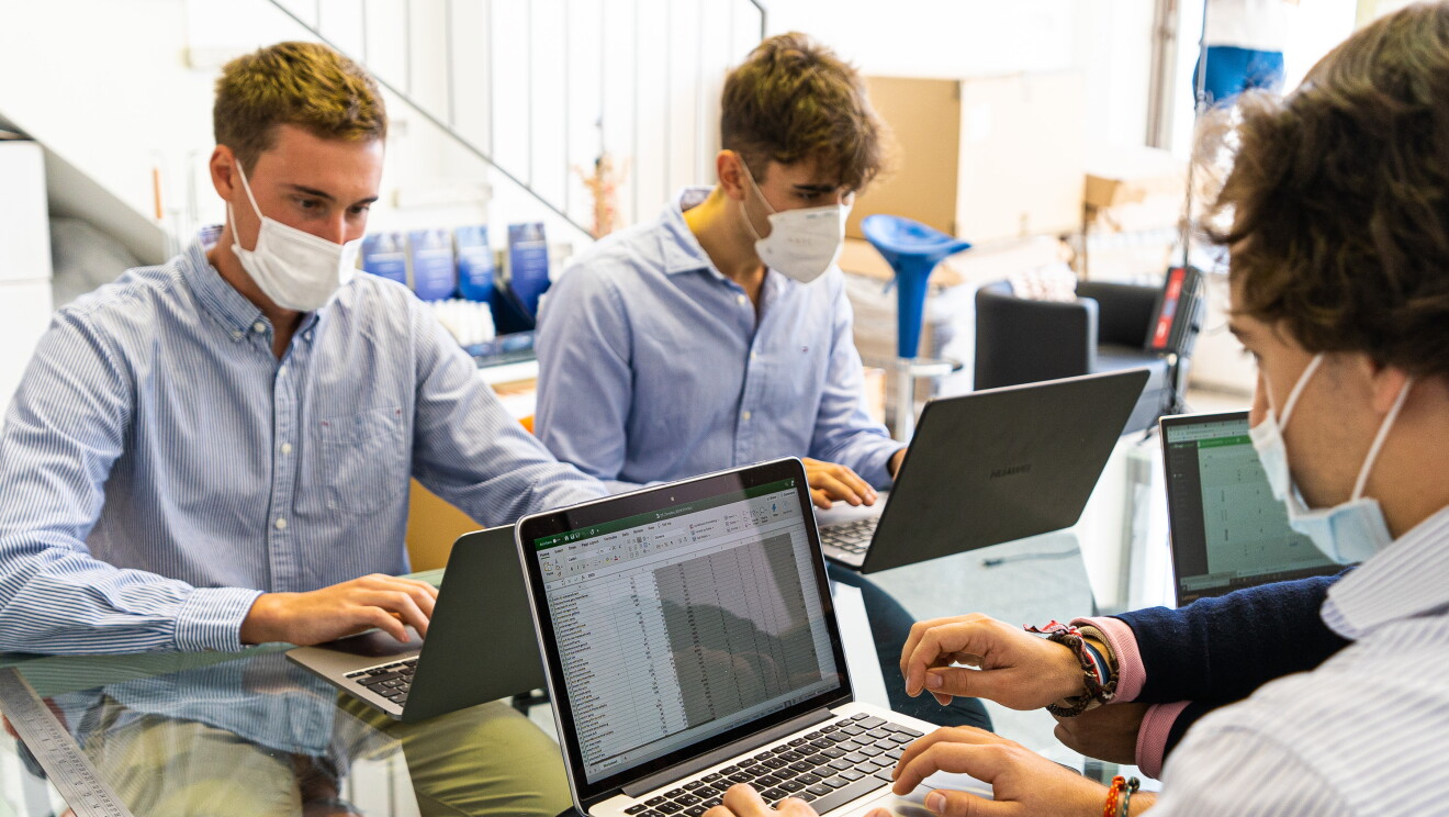 Javier Olivares, Jordi Cascant, Eloy Ribera y Iván Arenas forman EasyVal, el equipo ganador de la primera edición de Amazon Campus Challenge.  Eloy, Iván y Javier sentados en una mesa, cada uno con un ordenador. Se ven las manos de Jordi trabjando conjuntamente con Jordi. Eloy con una camisa azul de rayas y mascarilla. Iván también con camisa azul y mascarilla. Y Javier con camisa de rayas. Tiene un reloj en la mano izquierda y pulseras en la mano derecha. A su derecha aparecen las manos de Jordi, con una camisa rosa y un jersey azul. En la mano derecha tiene muchas pulselas. 
Los cuatro están sentados en una mesa de cristal. 
