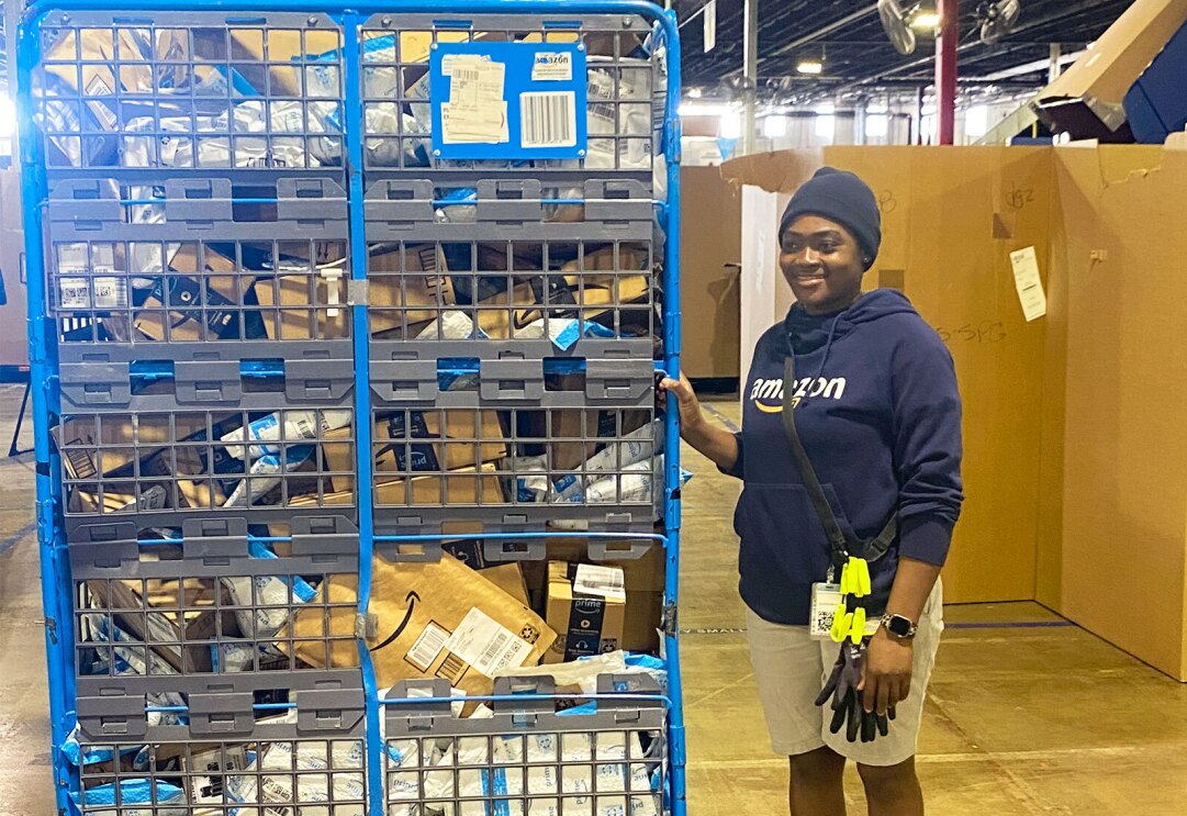 A photo of Amazon employee Abla Alassey standing inside a fulfillment center next to a large crate of Amazon packages.