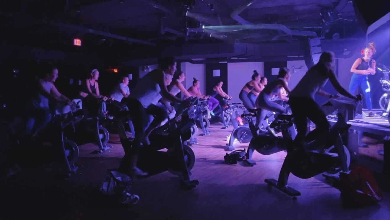 An image of a dark spin studio lit by a low, purple-tinted light. There are people riding stationary bikes and an instructor teaching at the front of the class.