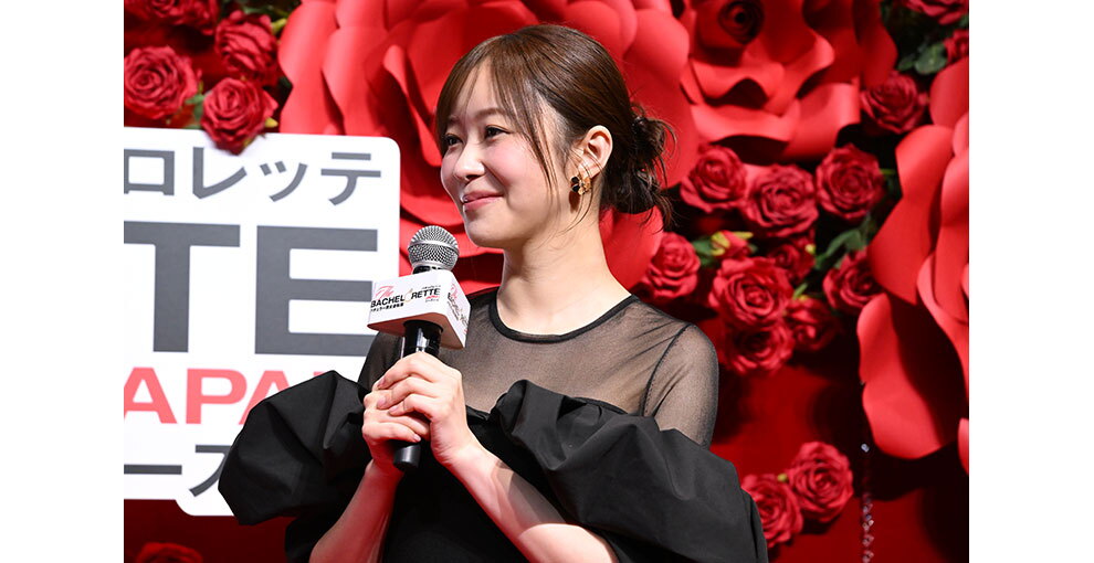 赤いバラの花の大きなオブジェと生花の赤いバラが飾られた赤い壁を背に、マイクを持って話す黒いドレス姿の女性。