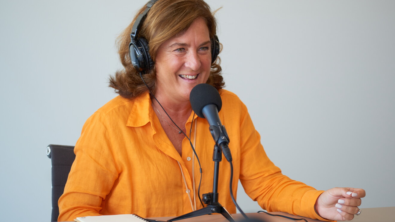 SARA BACEIREDO emprendedora y profesora del IE. sentada en una mesa delante de un micrófono y con unos auriculares puestos. Ella está mirando a un interlocutor que no se ve. Lleva el pelo suelto por el hombro  y color castaño- Una camisa de hilo de color naranja y un collar que no se ve el final poque queda por debajo de la mesa. Tiene los ojos azules muy claros y está muy morena de piel. Encima de la mesa tiene una libreta, un bolígrafo negro y una gafas morandas. 