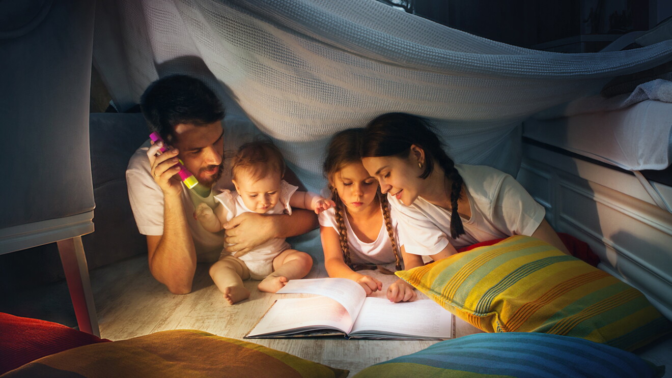 A family sits in a homemade fort, reading a book together by flashlight.
