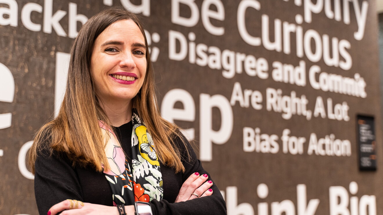 Lara Fajardo, Senior SW Development Manager Amazon Books. Llegó a Amazon hace un año. Plano por la cincuta de Lara. Con los brazos cruzados y sonriendo a cámara. Va con las u´ñas y los labios pintados de color rojo. Un jersey negro y un pañuelo de colores de flores estmapadas. De fondo una pared de las oficinas corporativas en Madrid con mensajes como: Bias for action, are right, a lot. 