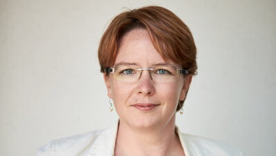 A woman smiles for a headshot photo in front of a white background while wearing a white blazer and glasses.