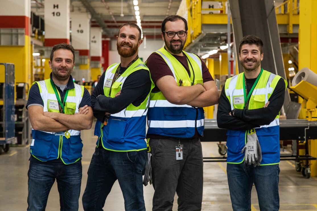 Quattro uomini con gilet ad alta visibilità e le braccia incrociate, guardano in camera e sorridono. Sono nel centro di distribuzione Amazon di Passo Corese.