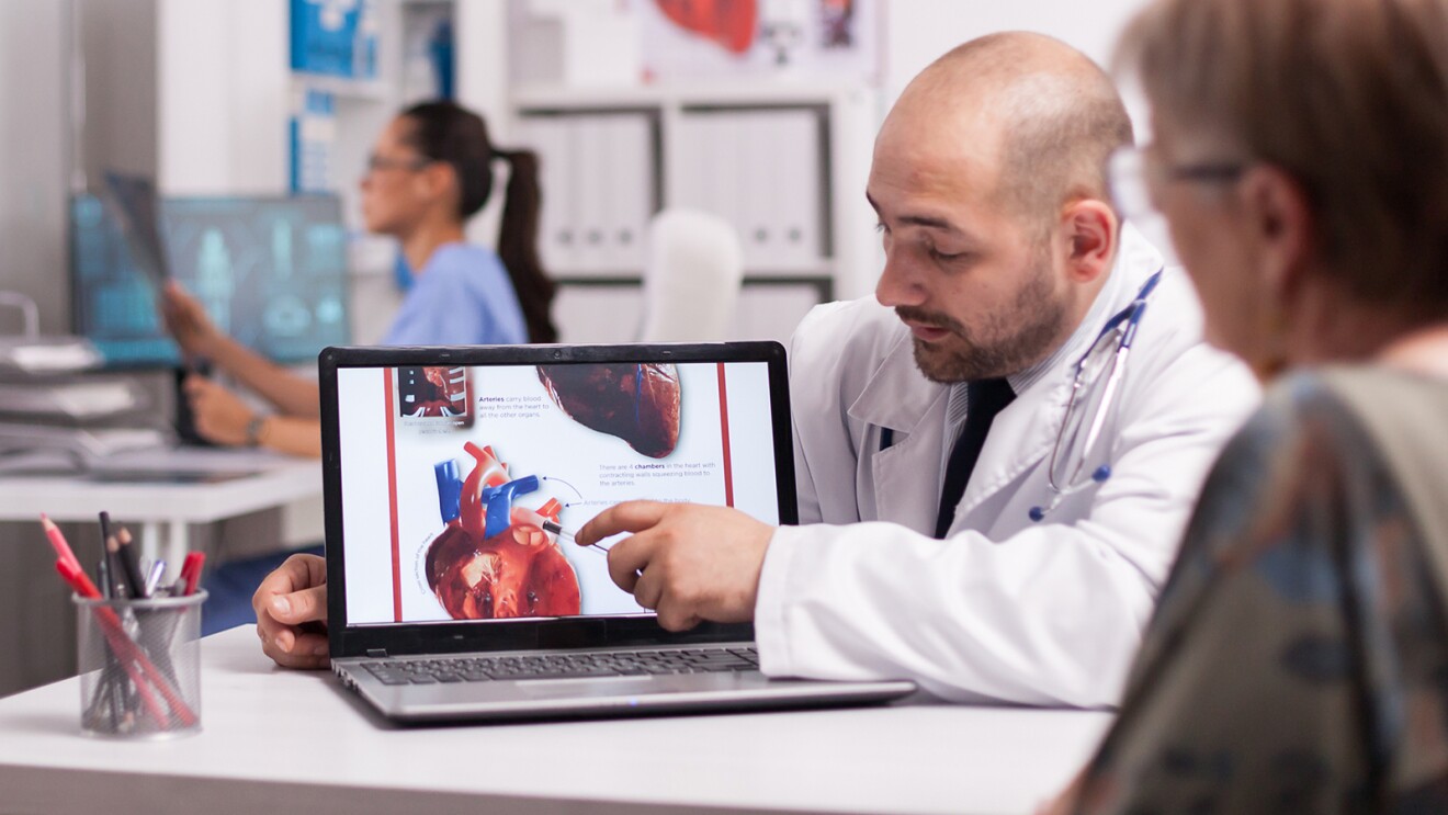 Médico mostra a imagem de um coração, na tela de um computador, a uma paciente idosa. É possível ver, ao fundo, uma médica analisando um raio-x