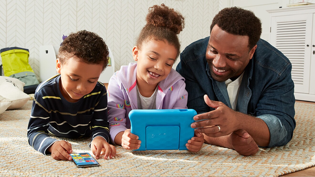 An image of a dad and two kids laying on a carpet smiling at their devices, including a tablet and a phone. 