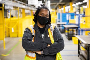 An Amazon employee stands in a fulfillment center with a mask on, looking at the camera.