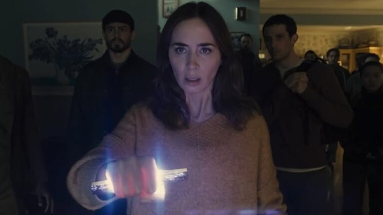 Women with a concerned expression stands in a group in a darkened room holding a light in her hands