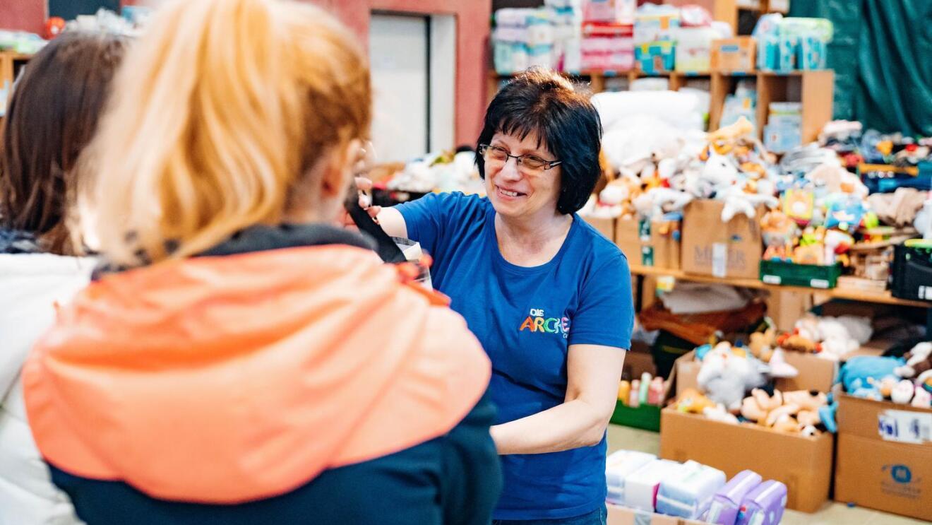 Ukraine Hilfe der Arche in Berlin Community Fund. Mitarbeiterin der Arche verteilt Spielzeugspenden an Kinder. Im Hintergrund sind weitere Kisten mit Spielzeug erkennbar.