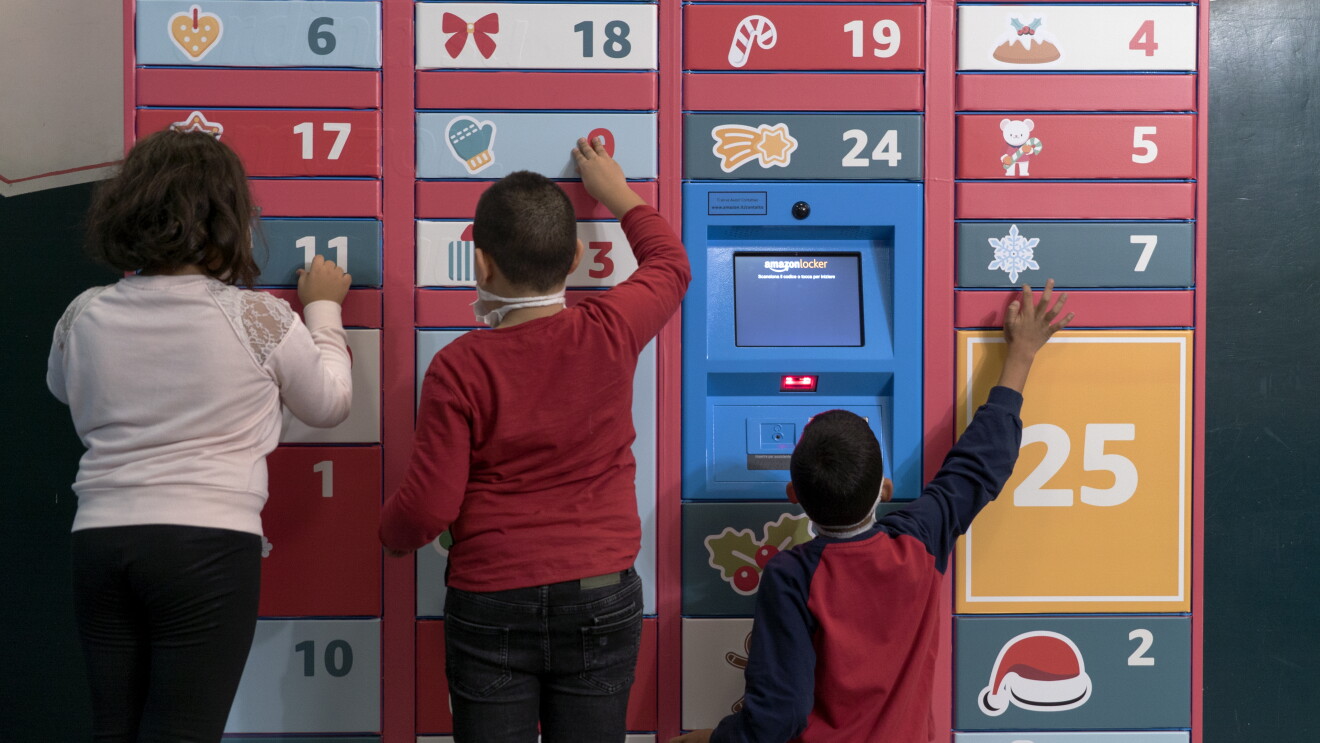 Locker Amazon ricoperto da adesivi natalizi su fondo rosso. Di fronte al Locker giocano 3 bambini, ripresi di spalle. 