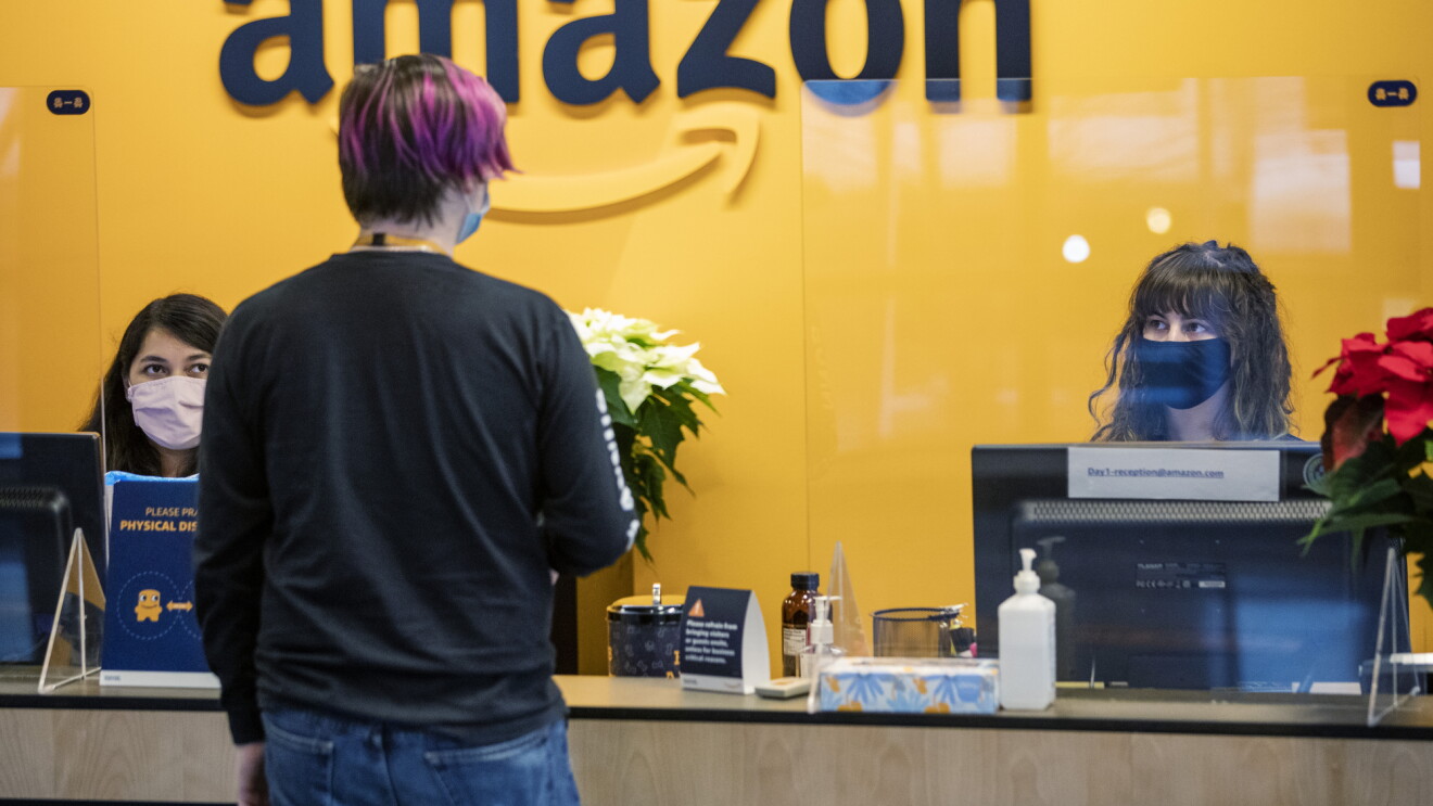 Amazon front desk employees speak with an Amazon employee. All are wearing masks, and there is a plexi divider between the receptionists and the employee. This photo was taken during the COVID-19 pandemic. 
