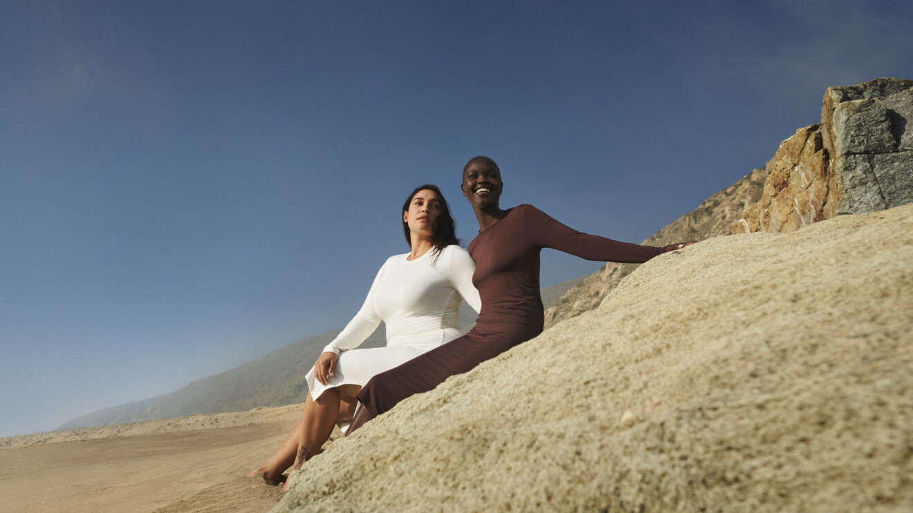 En una playa, apoyadas en una roca hay una chica negra y otra blanca. Las dos tienen uno de los brazos entrelazados y están apoyadas en una piedra con los pies en la arena. El vestido que llevan es blanco, la chica blanca y negro la chica negra. Es un vestido muy ajustado, de mandga larga, largo hasta después de la rodilla y cuello de camiseta. 