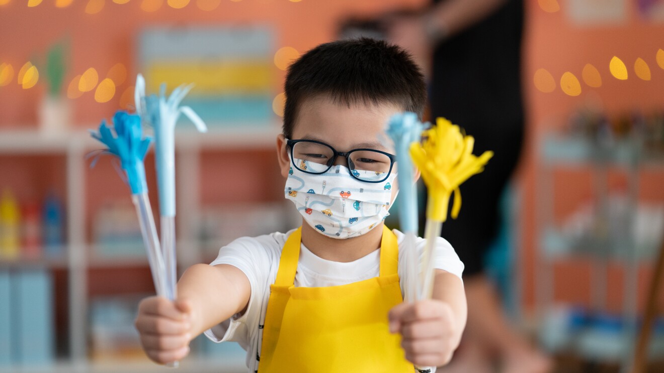 A young boy holding up his artwork, wearing a yellow apron