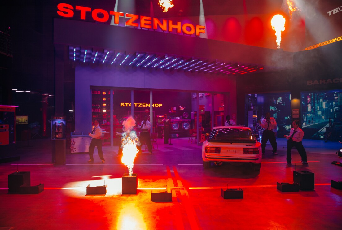 Episodenfoto von und mit Teddy Teclebrhan bei Prime Video. Man sieht eine Bühne mit brennender Fackel, ein Auto und die Bühnenaufschrift STOTZENHOF.