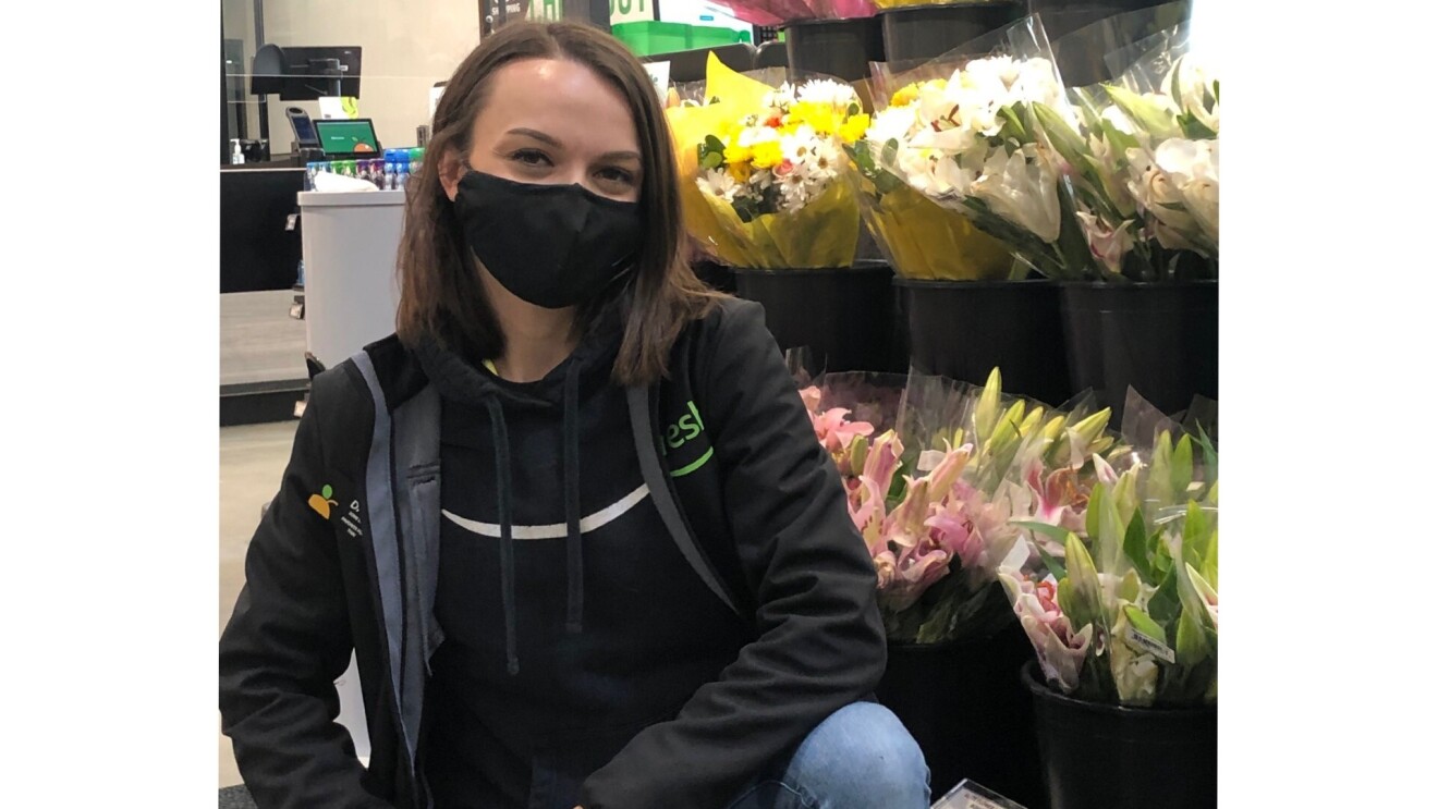 Darby wears an Amazon Fresh black jacket with a black hoodie underneath that has the Amazon logo on the front. She wears a face mask and has dark short hair. She smiles for the camera as she kneels down in front of flower bouquets at the new Amazon Fresh Store.