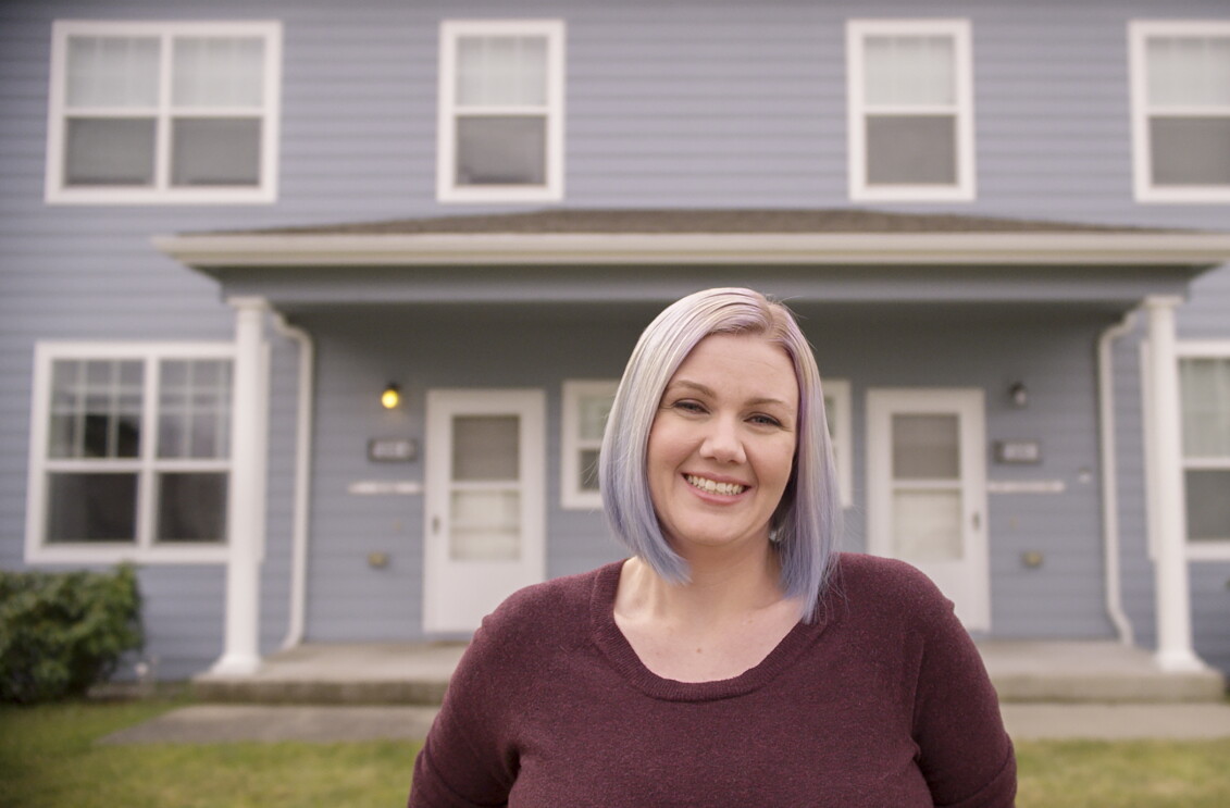 A woman stands in front of her home, smiling at the camera.