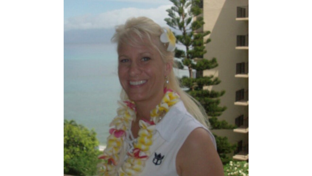 An image of a woman smiling for a photo on a balcony at a beachside resort. she is wearing a Hawaiian lei and a flower in her hair. The ocean and a tree are in the background.
