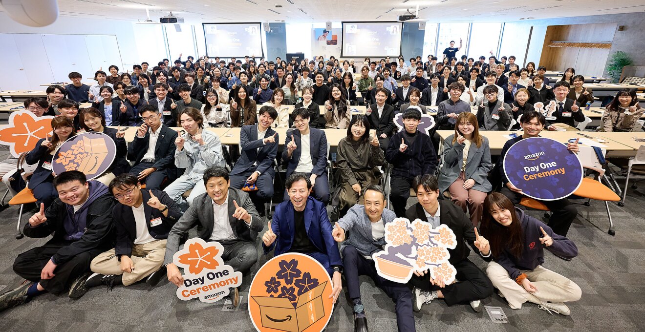 広いホールに集まり集合写真を撮る多数の人々。最前列の7人は床に座っている。2列目は椅子に座り、3列目以降は長机に向かって着席している。