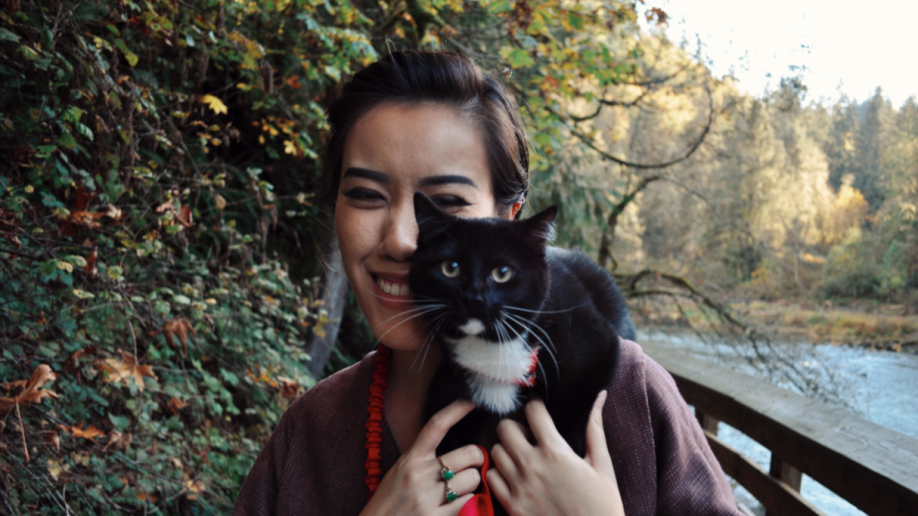 A woman wearing a purple coat, who is an Amazon intern, holding a black cat standing on a bridge next to a river.