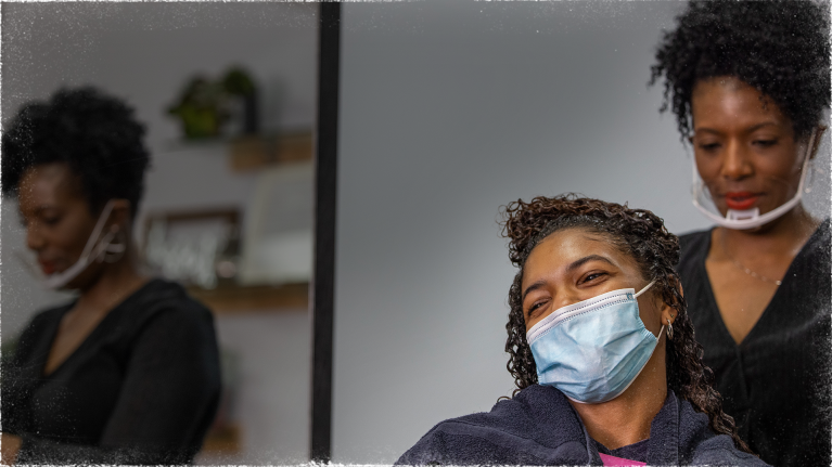 A woman wears a face mask in a stylist chair in a salon. Behind her, another woman wearing a mask is styling the first woman's hair.