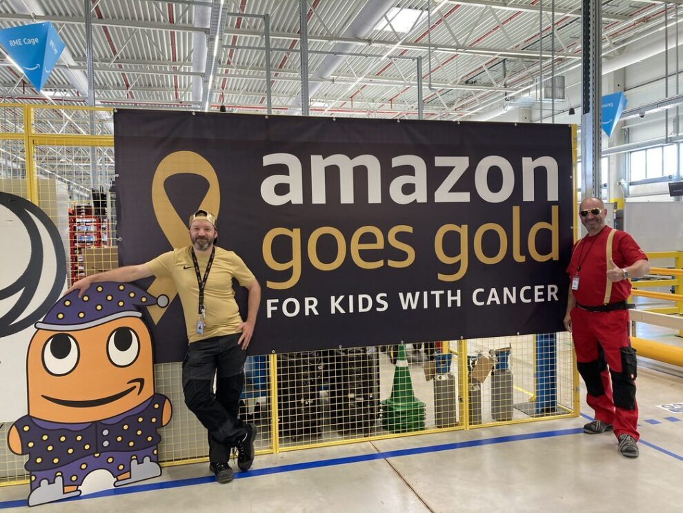 Zwei Amazon Mitarbeiter:innen vom Sortierzentum Halle stehen vor dem Amazon Goes Gold Banner und dem Amazon Maskottchen.