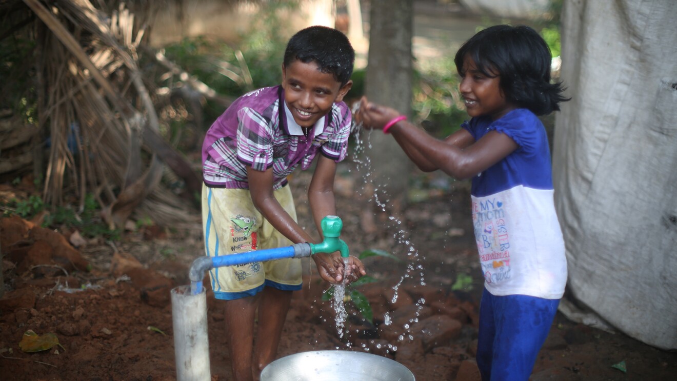 两个孩子站在水龙头旁的画面。他们面带微笑,伸出双臂捧起手中的水。