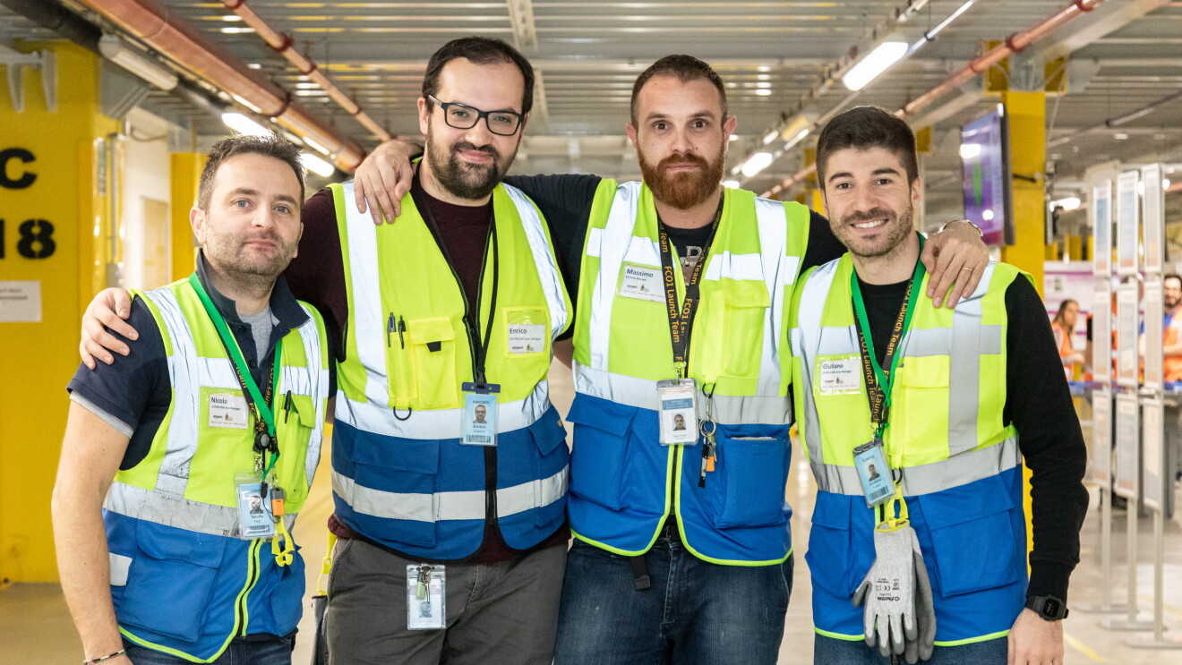 Quattro uomini guardano in camera e indossano gilet ad alta visibilità all'interno del centro di distribuzione Amazon di Passo Corese. 
