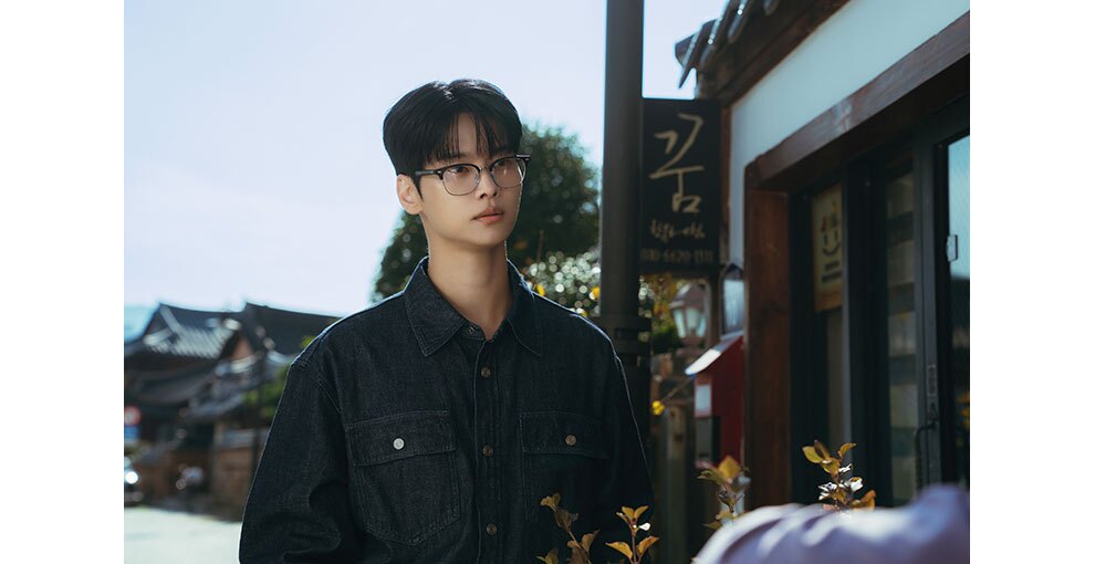 屋外の通りに立つ男性の上半身。右側の店には、ハングル文字の看板がかかっている。