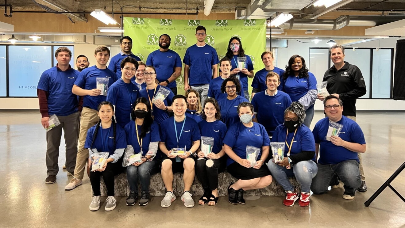 An image of AWS employees smiling for a group photo while holding hygiene kits they put together for people impacted in Ukraine. They are wearing matching blue shirts and there is a green background behind them that says "Clean the World."