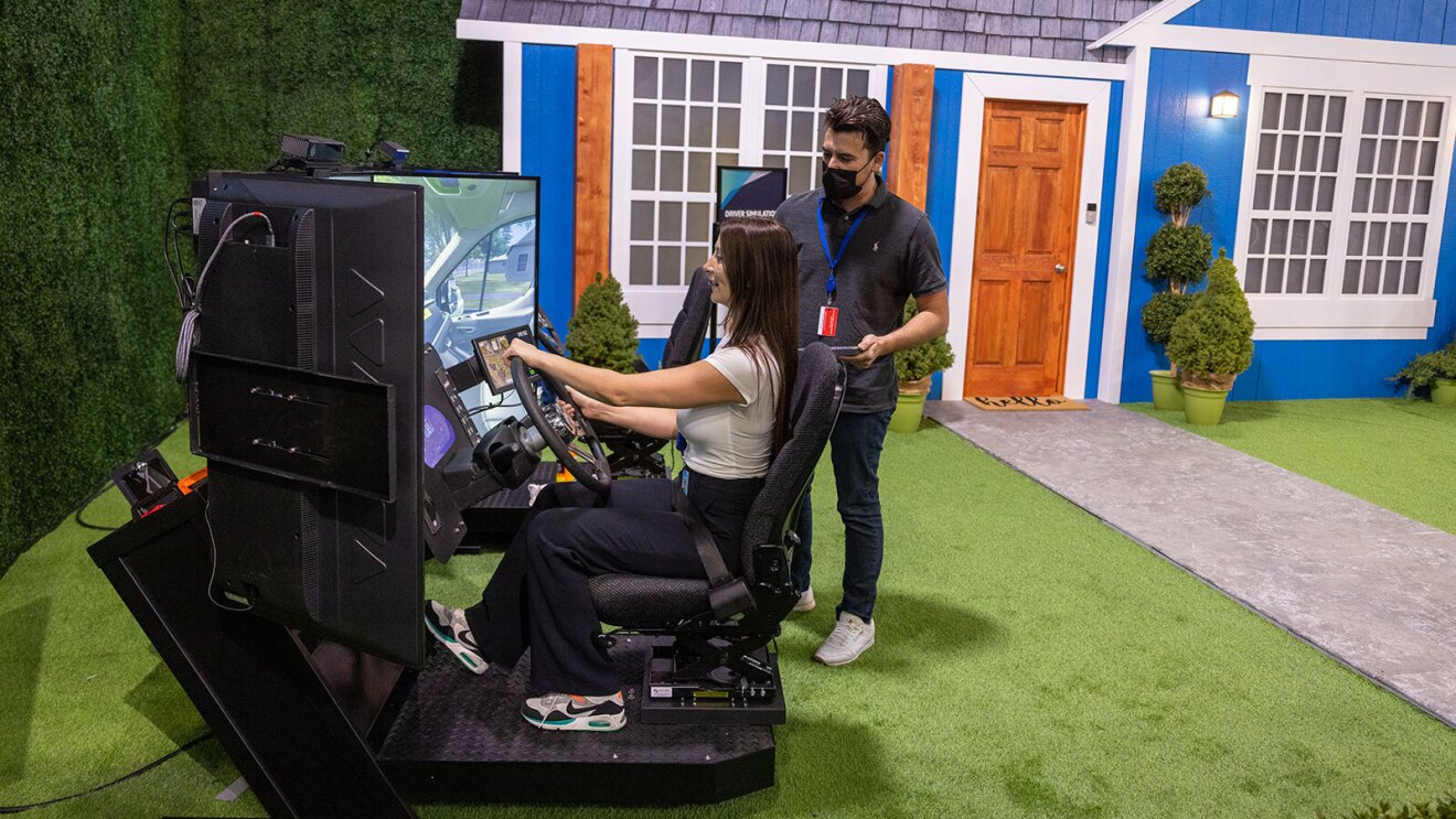 An image of a woman in a driving simulator. There is a driver's seat, a steering wheel, a dashboard, and three large screens in front of her. There is a man standing behind the simulator explaining how it works.