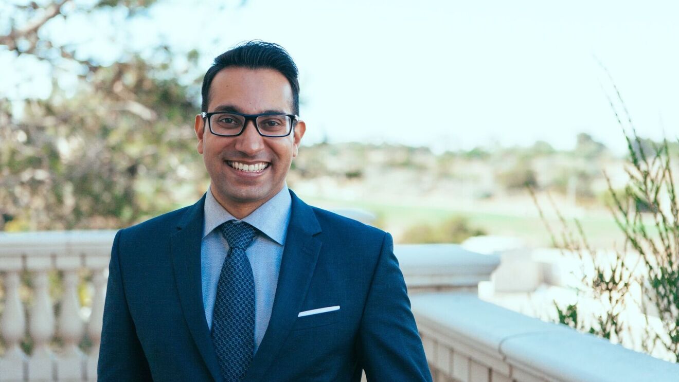Akash wears a navy suit as he stands outside and smiles for the camera. He has dark hair and wears black-framed eyeglasses.