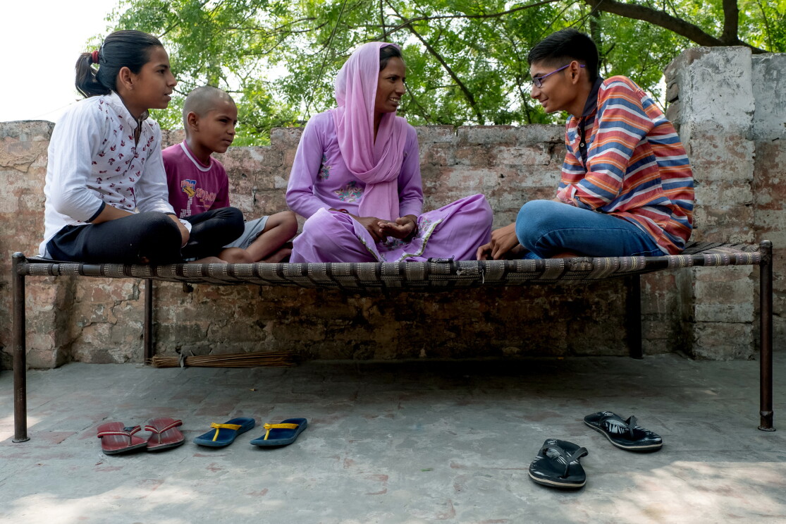 Three children sit outdoors on a bench with their mother. Three pairs of sandals are on the ground in front of the bench. A low brick wall and the leaves of a tree are in the background.