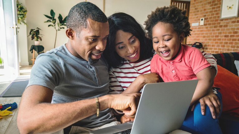 A family of three smile as they sit right next to each other on a couch looking at a laptop.