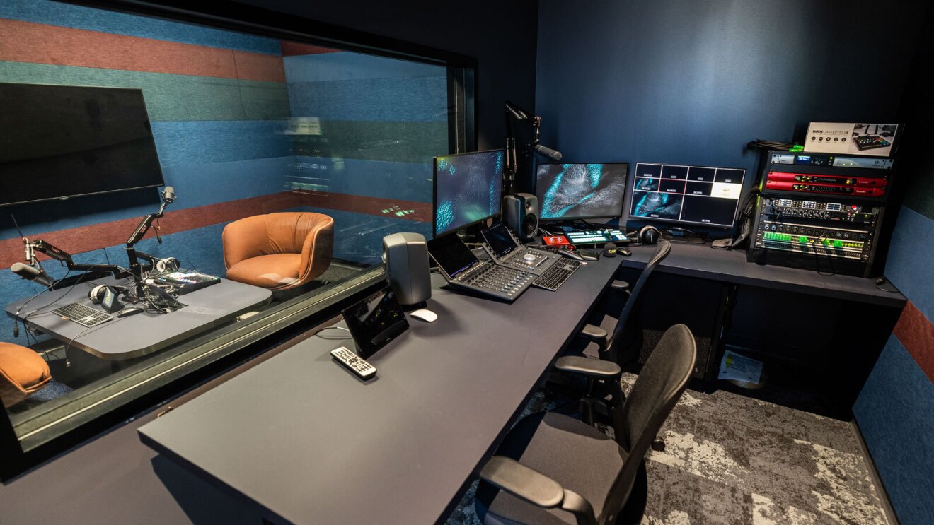 The view from the sound booth attached to one of Amazon Music's podcast studios in LA. There is a black table with chairs pulled up to it and several music controls and monitors on top. Through the glass, you see a podcast recording setup with chairs, microphones, and a TV.