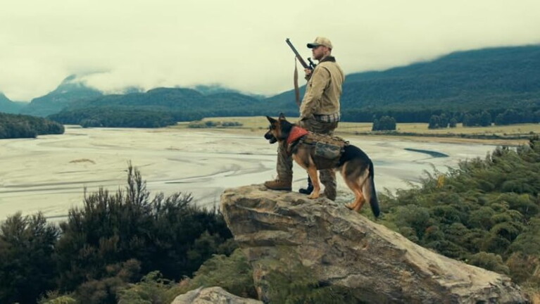 Hunter with rifle and German Shepherd overlooking a vast river valley from a rocky cliff