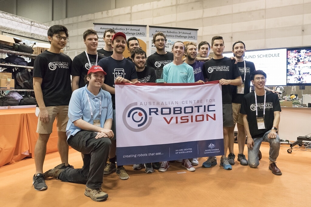 広いホールような屋内で集合写真を取る14人のチーム。前列が「ROBOTIC VISION」と書かれた旗を広げて持つ。2人を除いては揃いの黒いチームTシャツを着用。