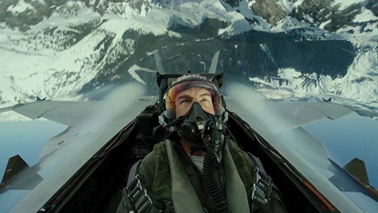 An image of the actor Tom Cruise in flight gear flying in the cockpit of a fighter jet in a scene from Top Gun: Maverick. The jet is upside down and there is a large, snowy mountain range below him.