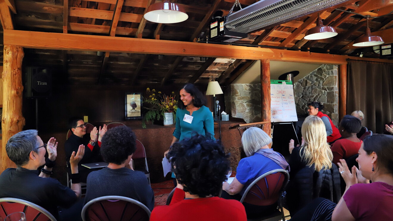 A woman stands in front of a crowd, who applauds from their seats. 
