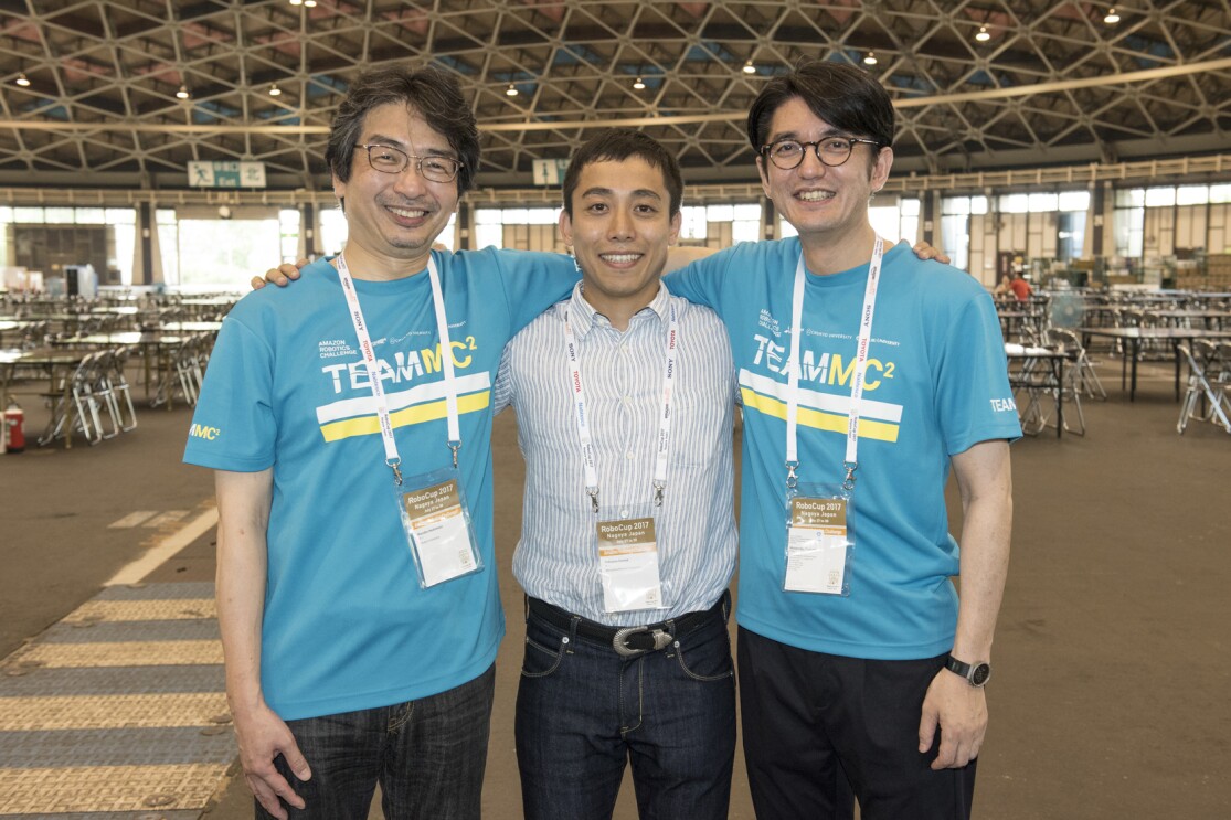 屋外ホールのような広い場所で肩を組んで並ぶ3人の男性。中央は青と白のストライプシャツ、左右の2人は水色の揃いのTシャツを着用。全員首から参加証のようなカードを下げている。