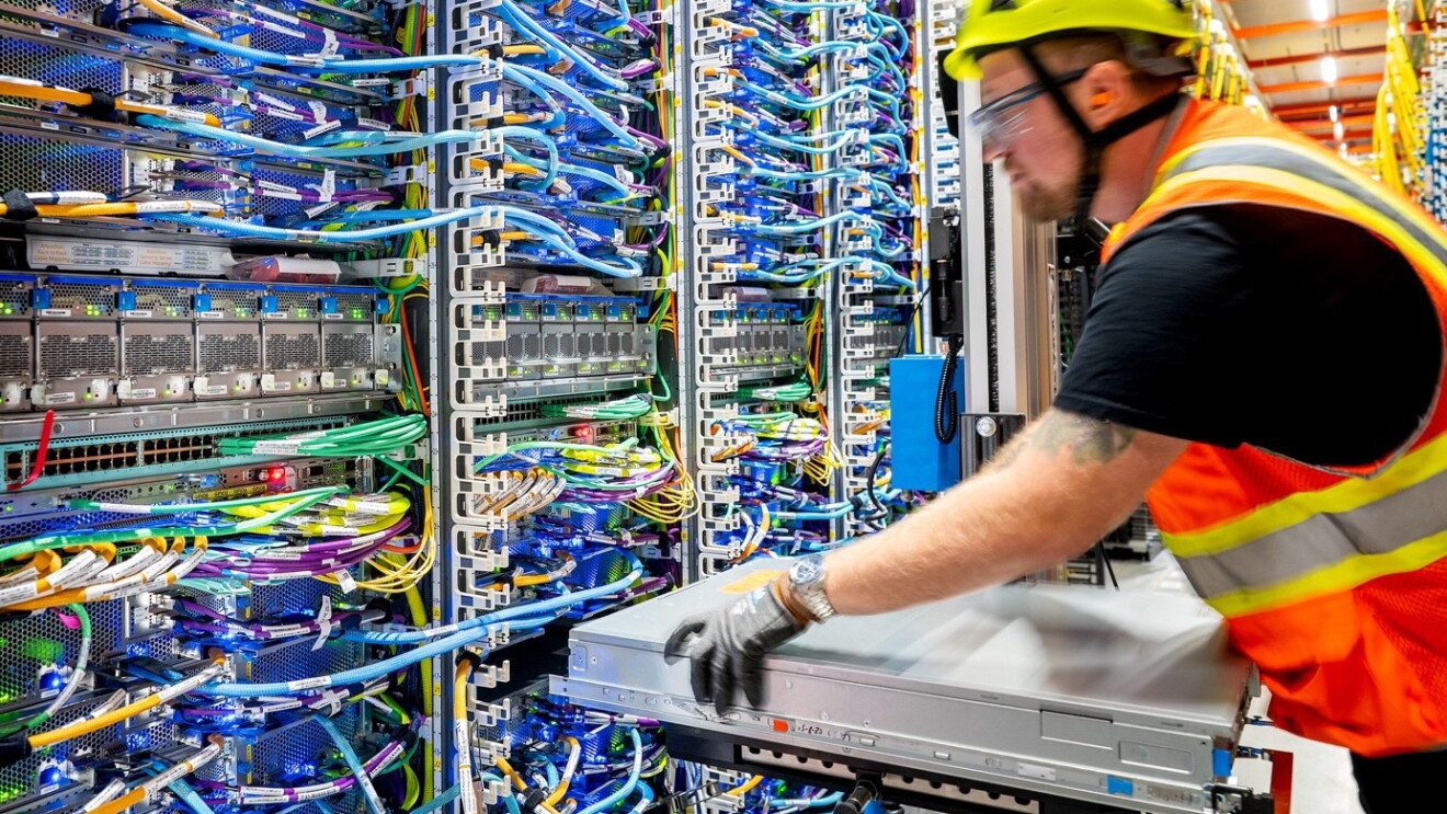 A man in an orange safety shirt and yellow safety hat in a data centre, working with data centre tower parts. 