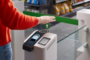 A person stands at a grocery entry gate with their hand suspended over a palm reading device. 