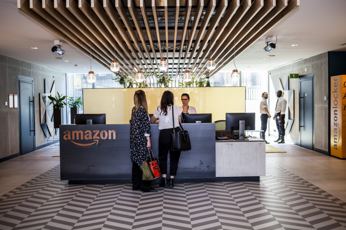 La reception degli uffici corporate di Amazon a Milano. Una donna dietroil bancone marrone, con il logo di Amazon sulla sinistra, accoglie due ospiti che rivolgono le spalle alla camera. In fondo, sulla destra, due guardie di sicurezza una di fronte all'altra parlano tra loro. 