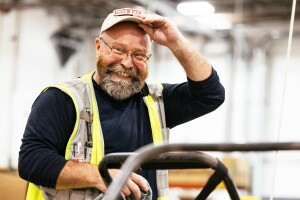 An operations employees wears a yello safety vest as he lifts up his hat and smiles.