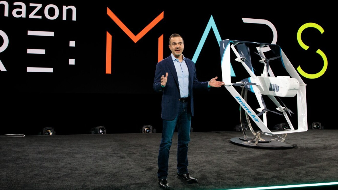 Mężczyzna w garniturze na scenie wydarzenia "Amazon Re:Mars", obok przykładowy prototyp Prime Air.
