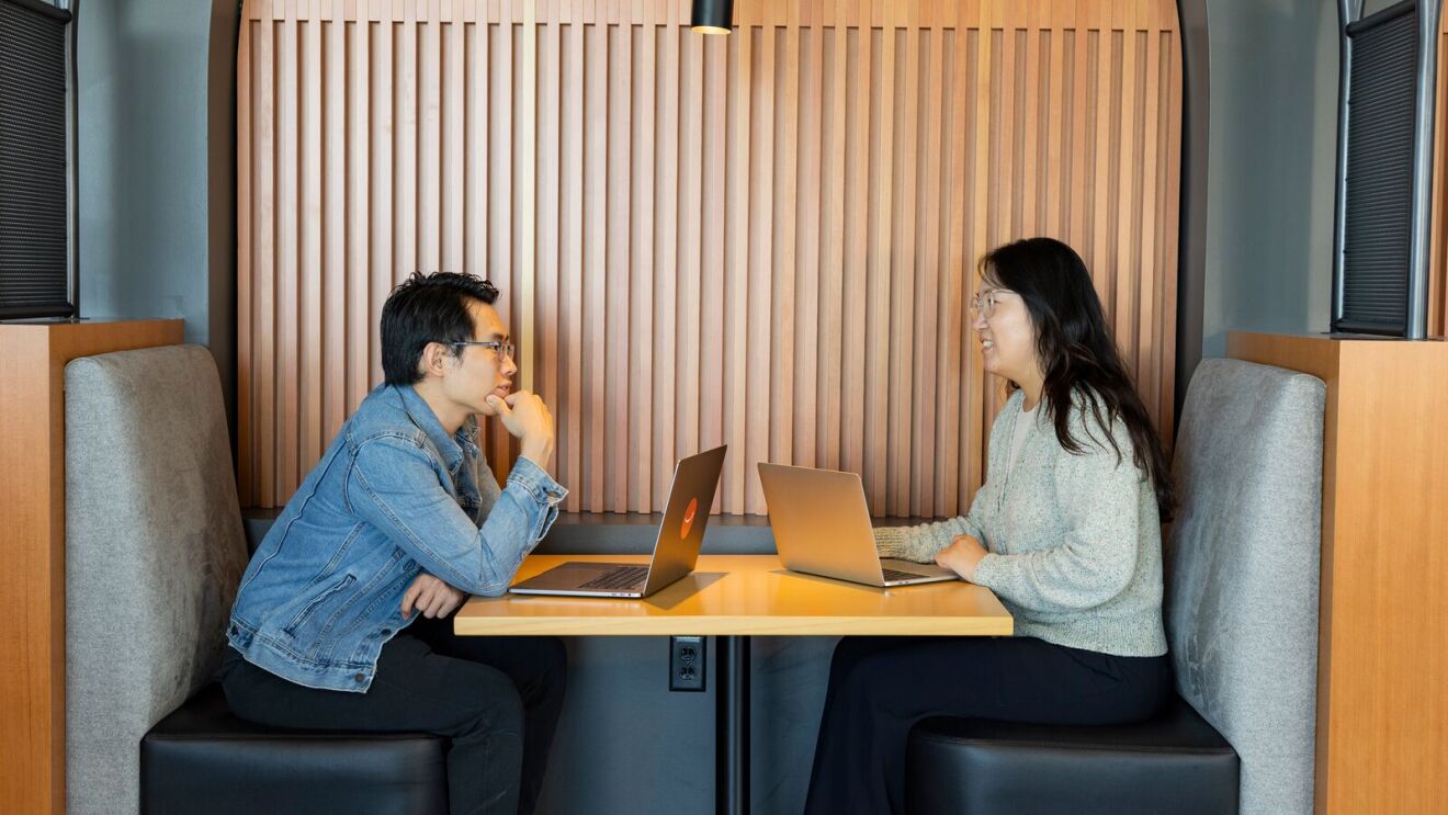A man and a woman chatting at a table with laptops.