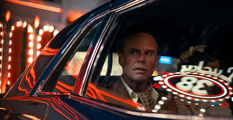 自動車の後部座席に座った男性のバストアップ。車の背後に明るいネオンが光り、赤いライトが車体と窓ガラスに反射している。男性の隣にも誰か座っているが、顔はネオンの反射で隠れて見えない。