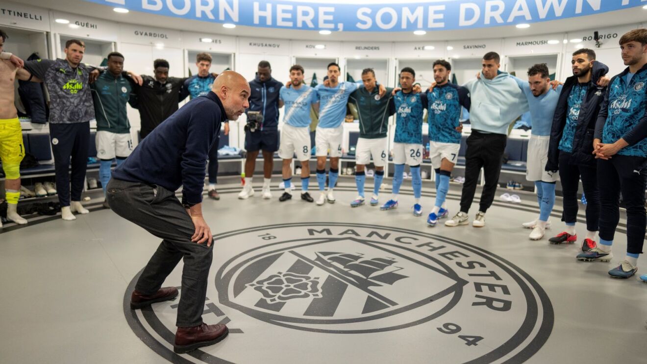 Pep Guardiola stands in the Man City dressing room, giving his players a team talk 